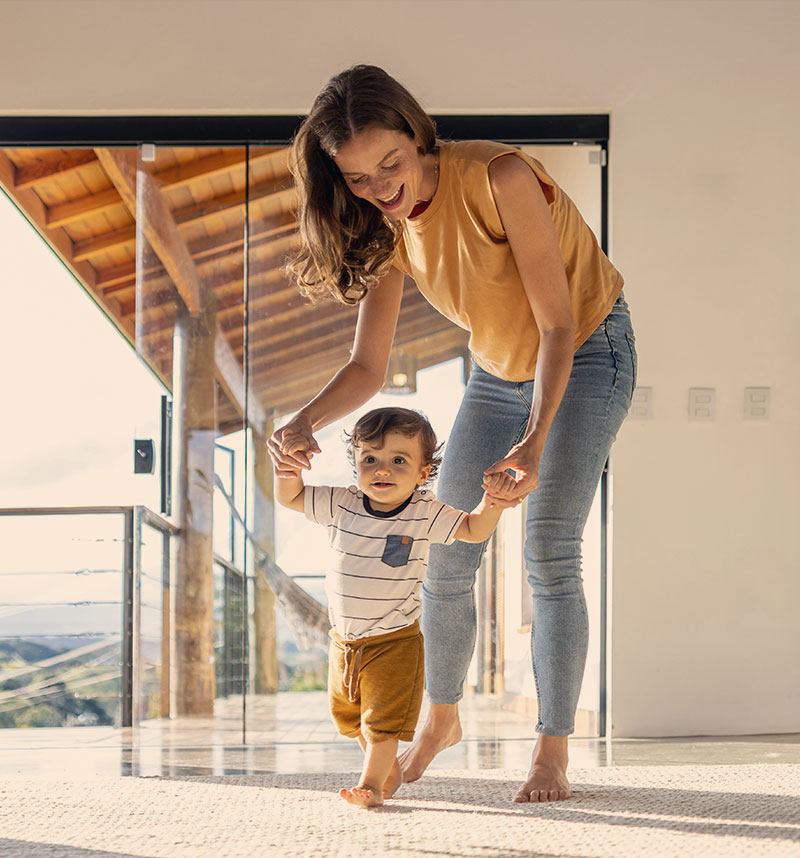Joyful mother assisting her toddler's first steps in a bright, sunlit home with modern architecture, conveying a sense of family support and growth.